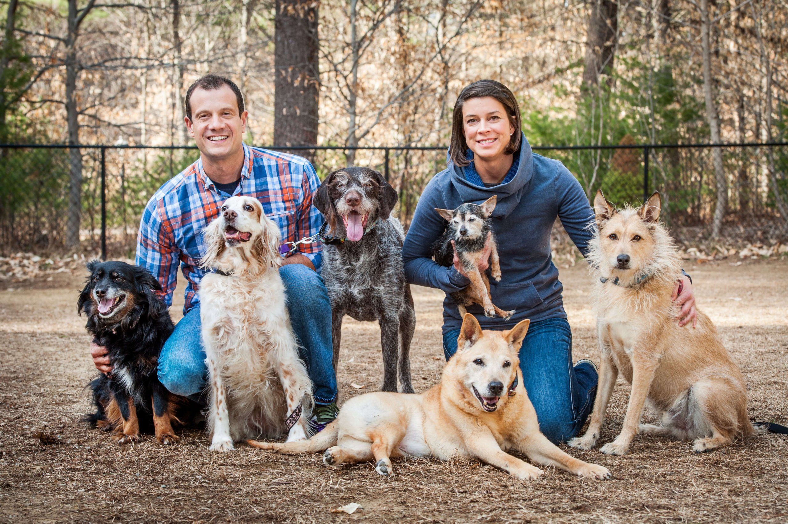a man and a woman sitting with 6 dogs outdoors