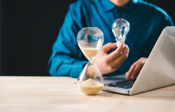 Hourglass time sand concept shows business idea emerging, glass timer countdown approaching, person holds lightbulb, creating innovation while working on laptop, developing new strategies.