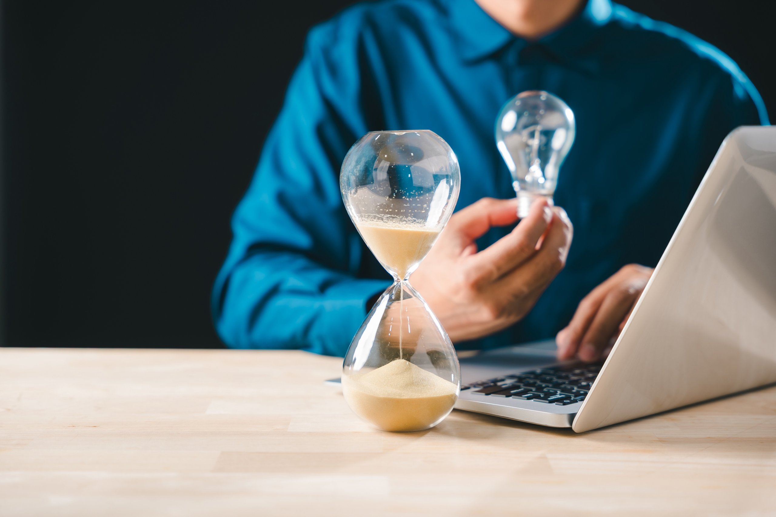 Hourglass time sand concept shows business idea emerging, glass timer countdown approaching, person holds lightbulb, creating innovation while working on laptop, developing new strategies.